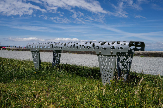 Reliance Foundry - A backless silver bench with leafy cutouts overlooks the ocean and a brilliant blue sky beside a seawall path